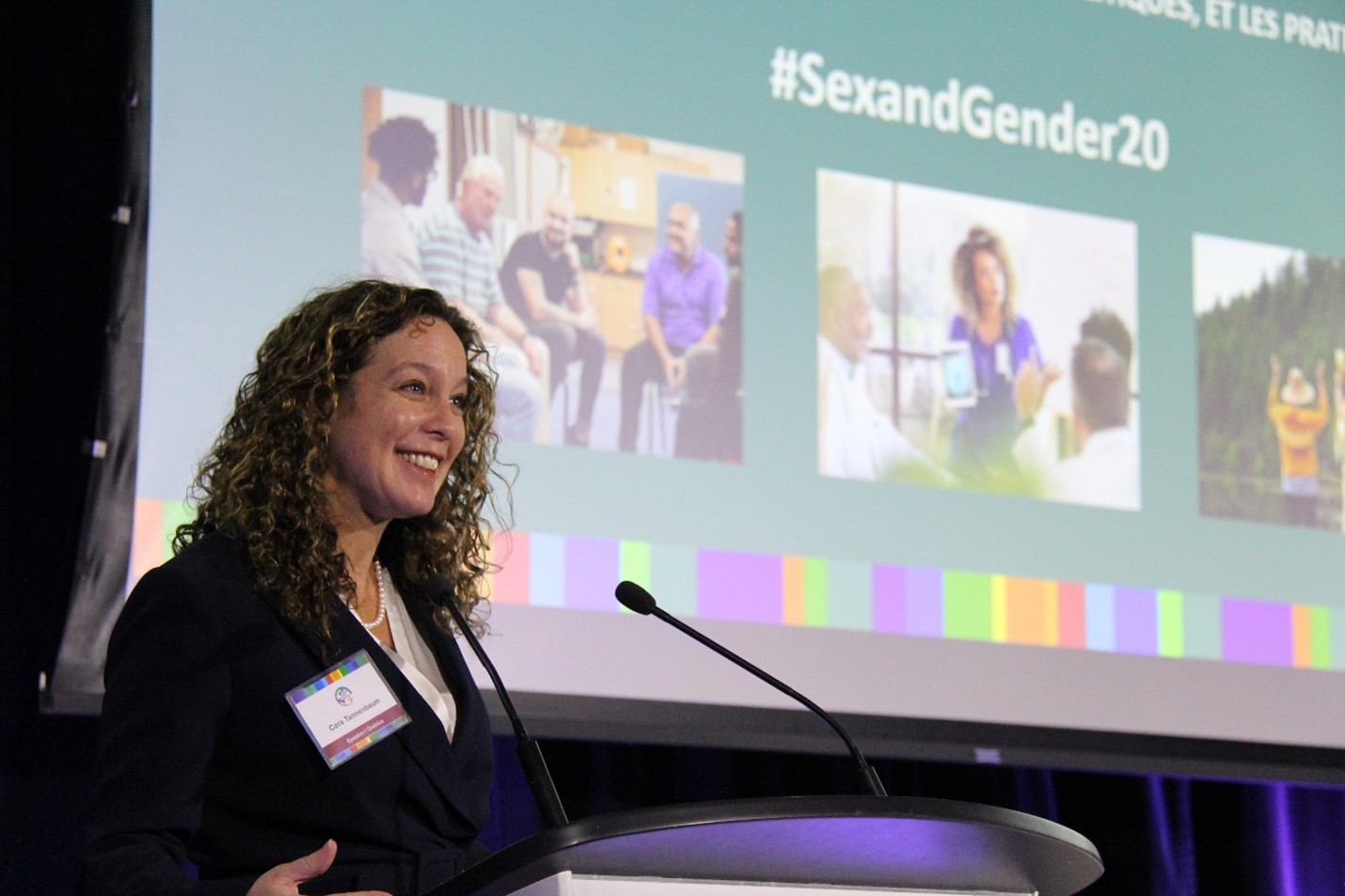 La Dre Cara Tannenbaum, debout sur un podium, regarde en avant et sourit. Derrière elle, un écran affiche des images colorées et le texte suivant : #SexAndGender20.